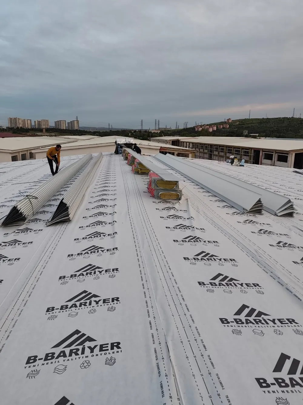 White B-Bariyer-printed roof membrane laid across an industrial hall roof, with metal sheet profiles and insulation packs on top and an installer at work